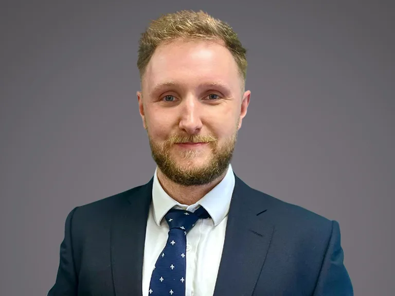Portrait of a person with short light-brown hair and beard, wearing a navy suit, white shirt and patterned blue tie, against a grey background.