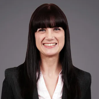 Person with long dark hair and blunt bangs, smiling broadly, wearing a white blouse and dark blazer, against a grey studio background.