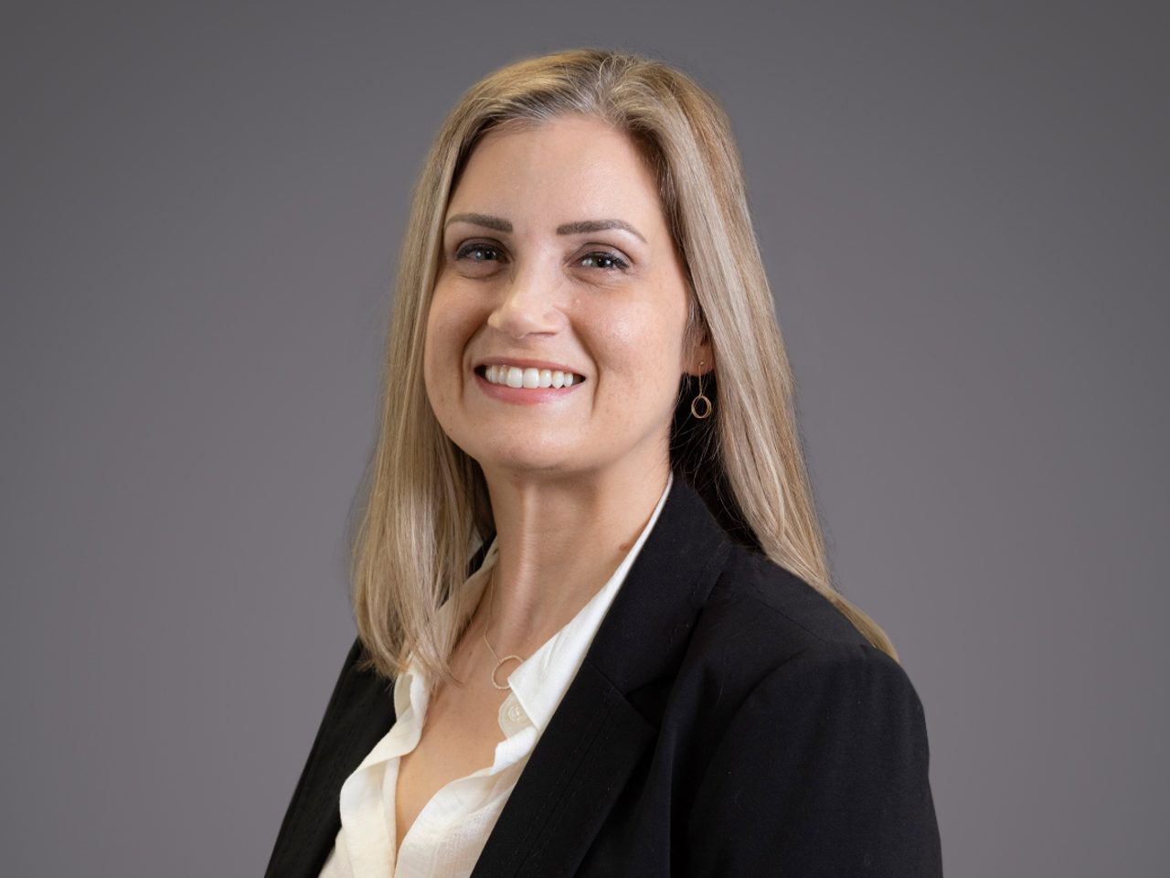 Professional portrait of a person with shoulder-length blonde hair, wearing a black blazer and white blouse, smiling against a grey backdrop.