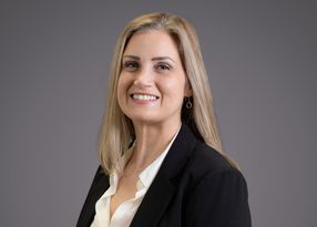 Professional portrait of a person with shoulder-length blonde hair, wearing a black blazer and white blouse, smiling against a grey backdrop.