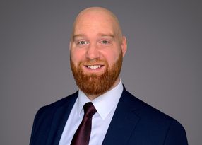Portrait of a bald person with a red beard, wearing a navy suit, white shirt, and burgundy tie, smiling against a gray backdrop.