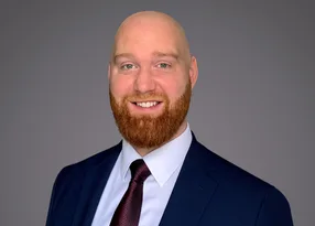 Head-and-shoulders portrait of a bald person with a red beard, wearing a navy suit, white shirt and burgundy tie, smiling against a grey background.