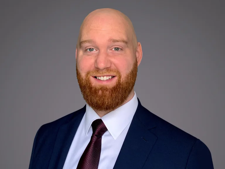 Head-and-shoulders portrait of a bald person with a red beard, wearing a navy suit, white shirt and burgundy tie, smiling against a grey background.