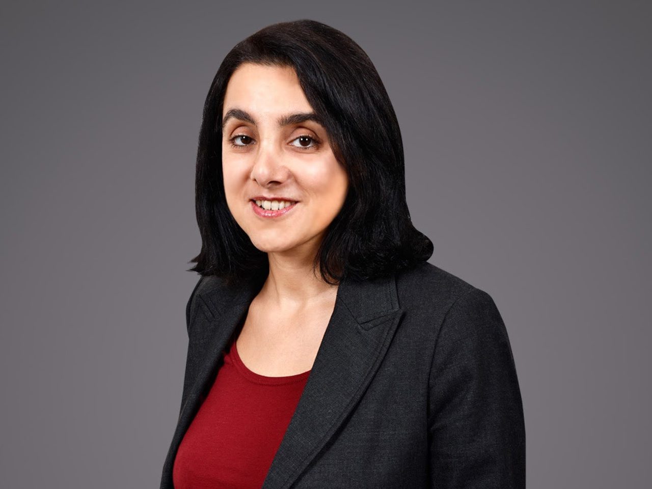 Portrait d'une personne aux cheveux noirs, souriante, portant blazer gris foncé et haut rouge, devant un fond gris uni.