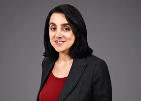 Portrait of a person with shoulder-length black hair, wearing a dark blazer over a red top, smiling against a grey studio backdrop.