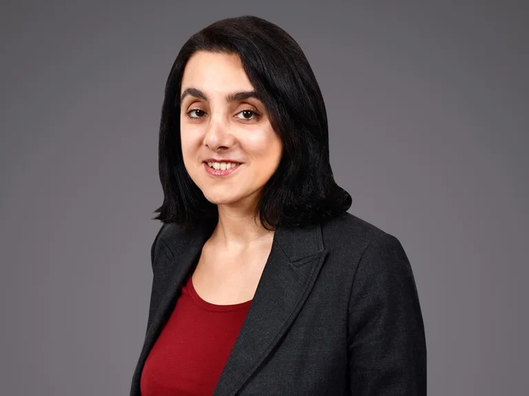 Portrait of a person with shoulder-length dark hair, wearing a dark blazer over a red top, smiling against a neutral gray background.