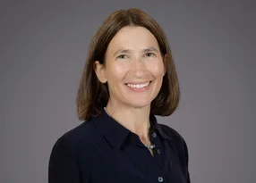 A smiling woman with shoulder-length brown hair, wearing a dark blue shirt and pearl necklace, poses against a gray background.