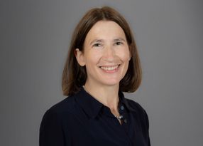 Portrait of a smiling person with shoulder-length brown hair, wearing a dark shirt and a pearl necklace, against a grey backdrop.