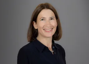 Portrait of a smiling person with shoulder-length brown hair, wearing a dark shirt and a pearl necklace, against a grey backdrop.