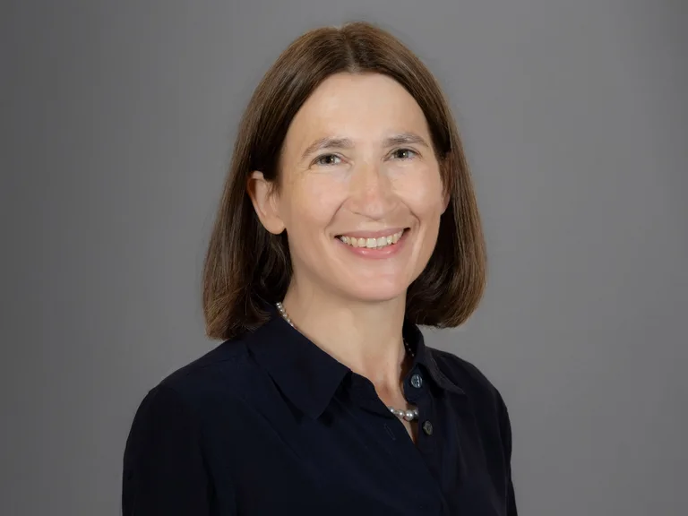 Portrait of a smiling person with shoulder-length brown hair, wearing a dark shirt and a pearl necklace, against a grey backdrop.