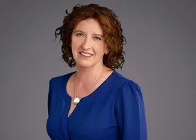 Person with short curly brown hair, wearing a royal blue blouse and a silver necklace with a white pendant, smiling against a neutral grey background.