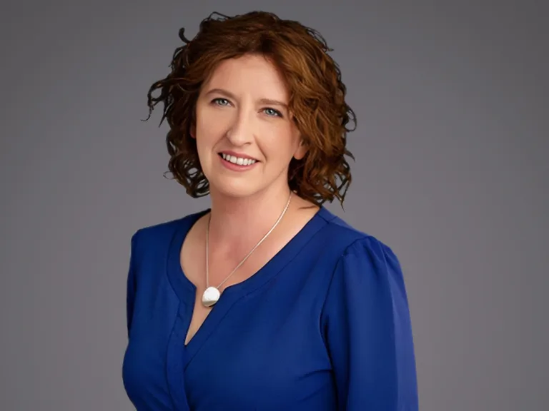 Person with short curly brown hair, wearing a royal blue blouse and a silver necklace with a white pendant, smiling against a neutral grey background.