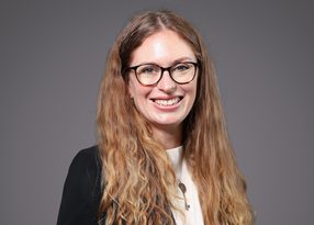 Smiling person with long wavy light brown hair, wearing glasses, a white top and black blazer, against a grey backdrop.