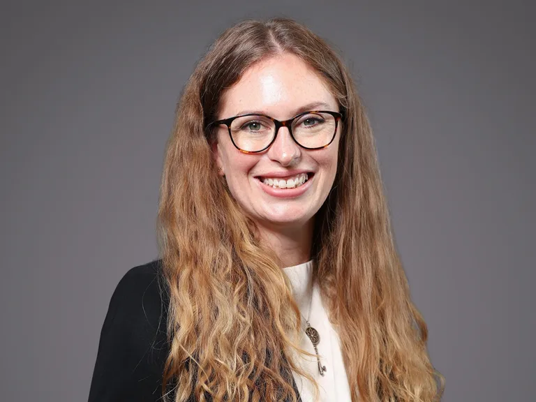 Smiling person with long wavy light brown hair, wearing glasses, a white top and black blazer, against a grey backdrop.