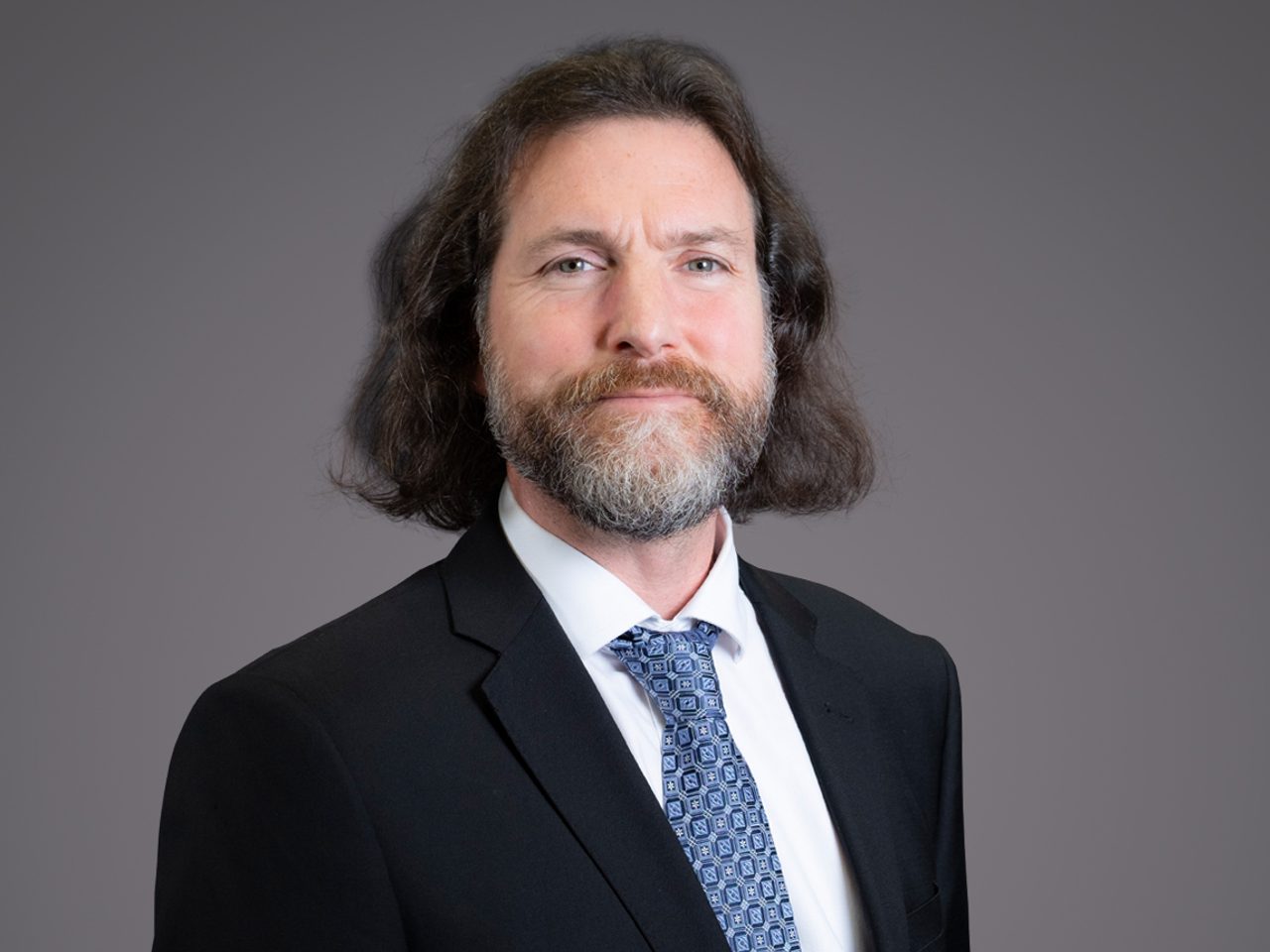 Portrait of a person with shoulder-length hair and a beard, dressed in a dark suit, white shirt, and blue patterned tie, against a grey backdrop.