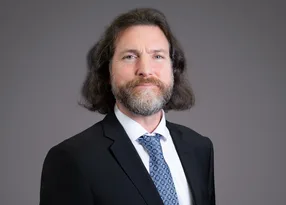 Portrait of a person with shoulder-length hair and a beard, dressed in a dark suit, white shirt, and blue patterned tie, against a grey backdrop.