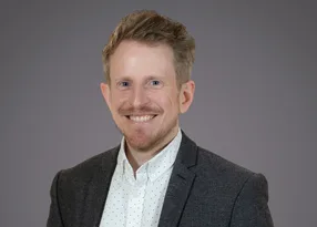 A young man with short, light brown hair, blue eyes, and a beard, wearing a patterned blazer and white shirt with small black dots, smiling against a gray background.