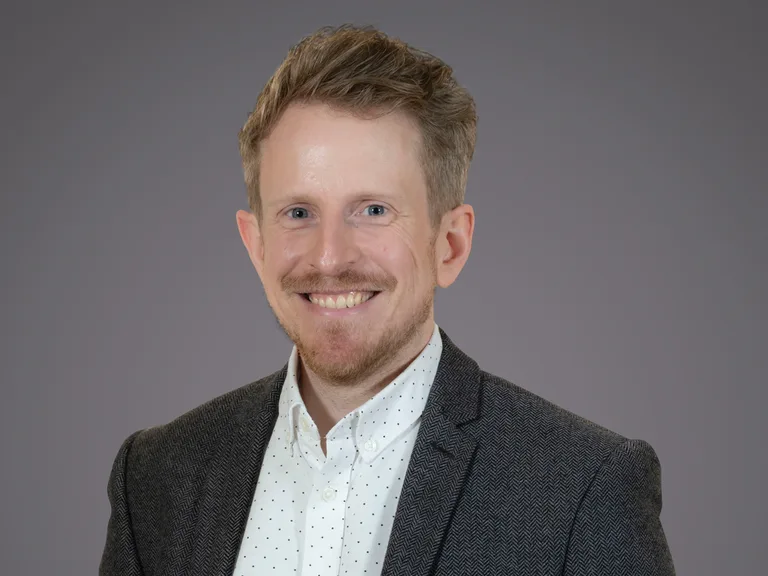 Portrait of a person with short light-brown hair, light stubble, smiling, in a dark blazer over a white polka-dot shirt against a gray background.