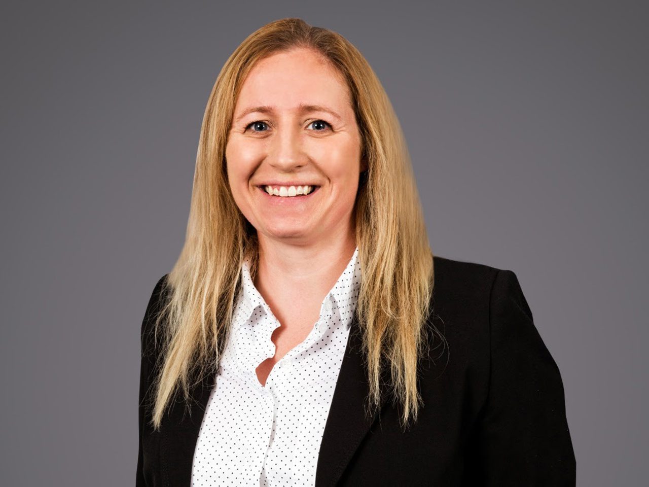 Portrait of a person with long blonde hair, wearing a black blazer over a white polka-dot blouse, smiling against a grey studio backdrop.