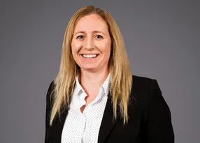 Portrait of a person with long blonde hair, wearing a black blazer over a white polka-dot blouse, smiling against a grey studio backdrop.
