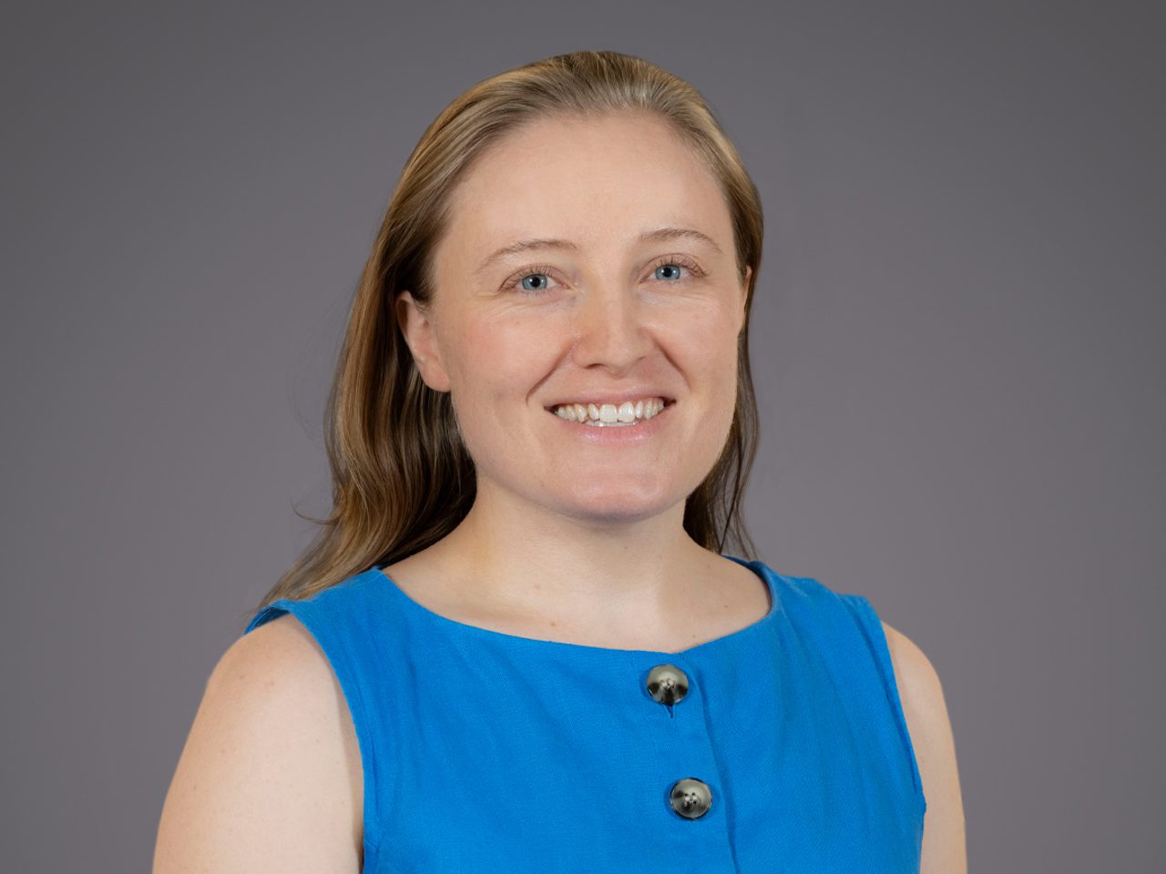 Portrait of a person with light brown hair and blue eyes, wearing a blue sleeveless top with two black buttons, smiling against a grey backdrop.