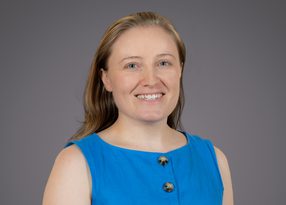 Portrait of a person with light brown hair and blue eyes, wearing a blue sleeveless top with two black buttons, smiling against a grey backdrop.