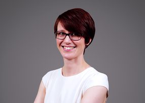 Portrait of a person with short dark red hair and glasses, wearing a white top, smiling at the camera against a grey background.