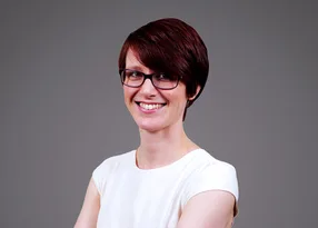 Portrait of a person with short dark red hair and glasses, wearing a white top, smiling at the camera against a grey background.