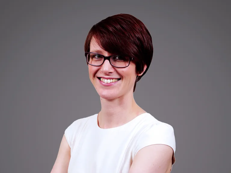 Portrait of a person with short dark red hair and glasses, wearing a white top, smiling at the camera against a grey background.