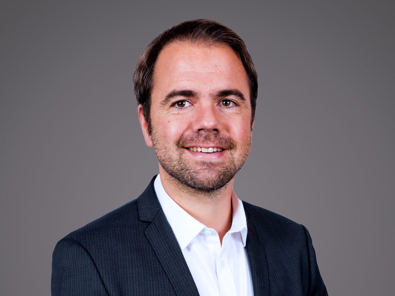 Portrait d'une personne souriante portant une chemise blanche et un blazer sombre à rayures fines, sur fond gris neutre.