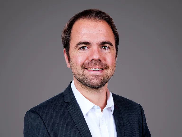 Portrait of a person with short dark hair and light stubble, wearing a dark pinstripe suit and white shirt, smiling against a grey studio backdrop.