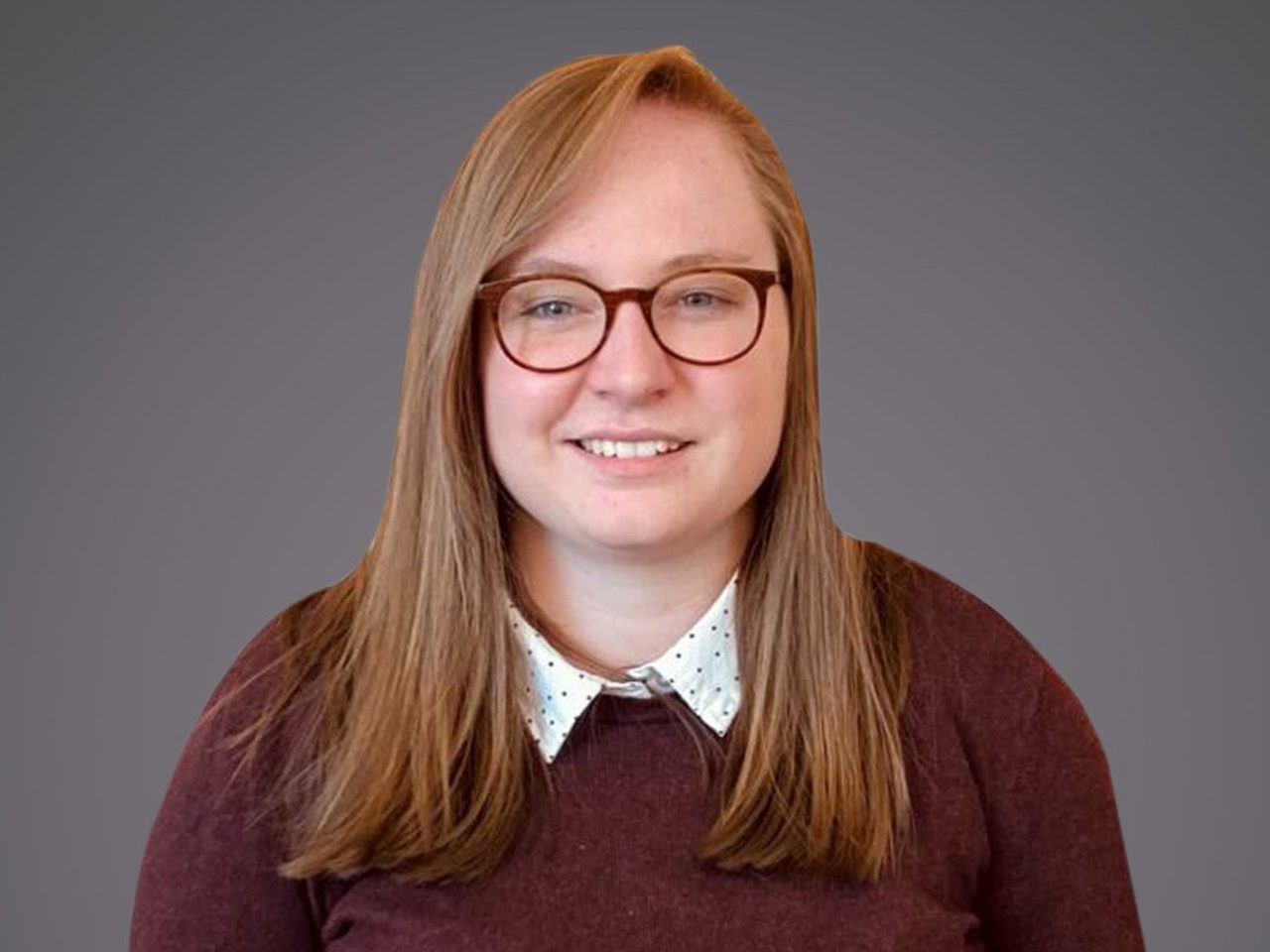 Person with long light brown hair and round glasses, smiling at the camera, wearing a white polka-dot shirt under a maroon sweater.