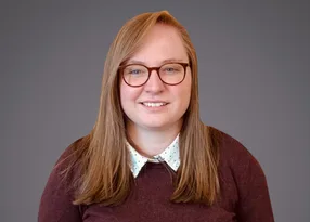 Person with long light brown hair and round glasses, smiling at the camera, wearing a white polka-dot shirt under a maroon sweater.