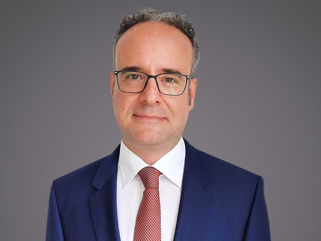 Portrait of a person with short grey hair and glasses, wearing a navy suit, white shirt and red tie, against a grey gradient background.