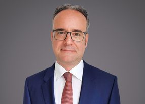 Portrait of a person with short grey hair and glasses, wearing a navy suit, white shirt and red tie, against a grey gradient background.