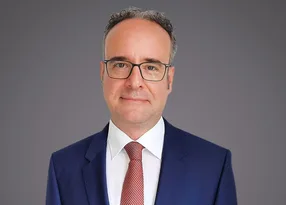 Portrait of a person with short grey hair and glasses, wearing a navy suit, white shirt and red tie, against a grey gradient background.