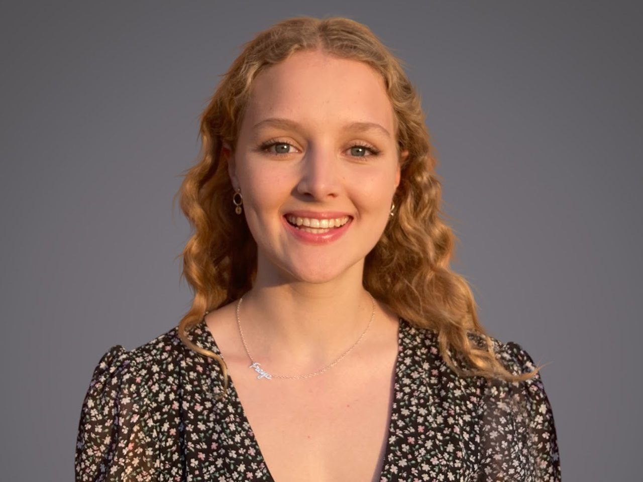 A smiling person with shoulder-length, wavy blond hair wearing a black floral-patterned blouse, a delicate necklace, and small hoop earrings, against a gray studio background.