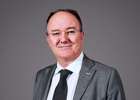 Portrait of a person in a grey suit, white shirt and black tie, wearing glasses, smiling at the camera against a grey backdrop.