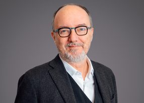 Portrait of a person with short grey beard and glasses, wearing a white shirt and dark blazer, against a plain grey background.