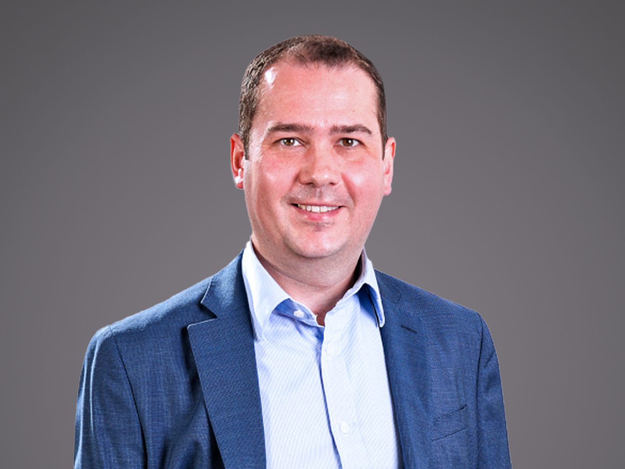 A person in a blue blazer and light blue shirt smiles at the camera against a neutral grey gradient background.