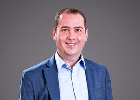 A person in a blue blazer and light blue shirt smiles at the camera against a neutral grey gradient background.