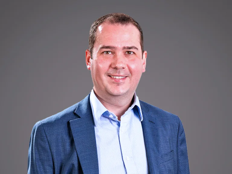 A person in a blue blazer and light blue shirt smiles at the camera against a neutral grey gradient background.