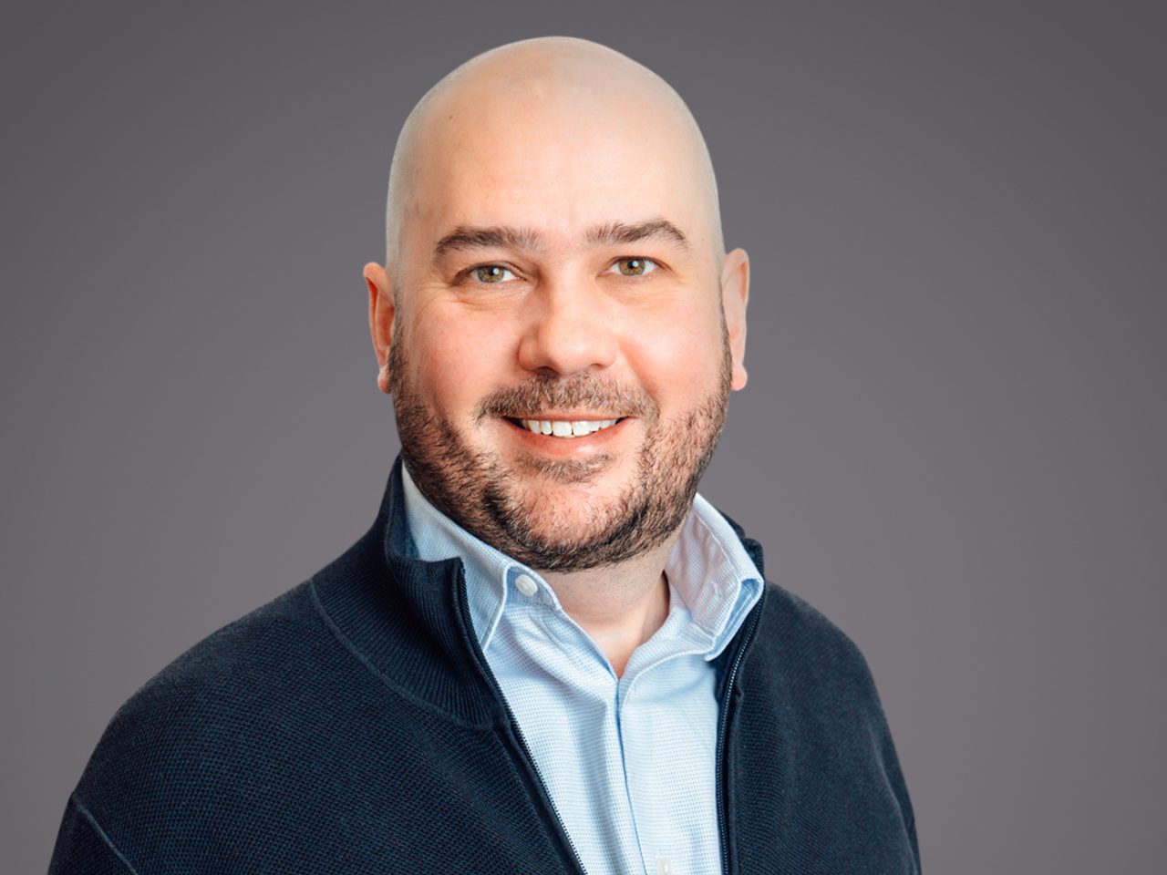 Head-and-shoulders portrait of a bald person with a short beard, wearing a light blue shirt under a dark zip sweater, smiling against a gray background.