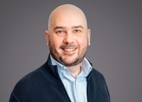 Bald person with a trimmed beard, wearing a light blue shirt under a dark navy zip-up sweater, smiling against a grey studio backdrop.