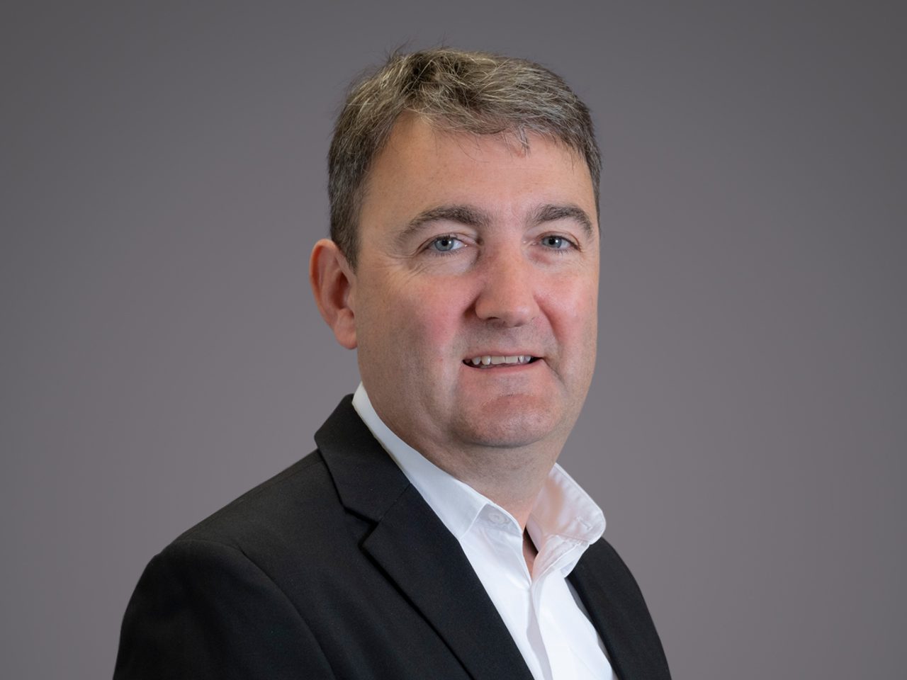 Portrait en buste d'une personne avec un léger sourire, veste noire et chemise blanche, fond gris neutre.