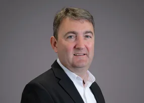 Portrait of a person wearing a dark blazer and white shirt, smiling slightly, against a neutral grey background.