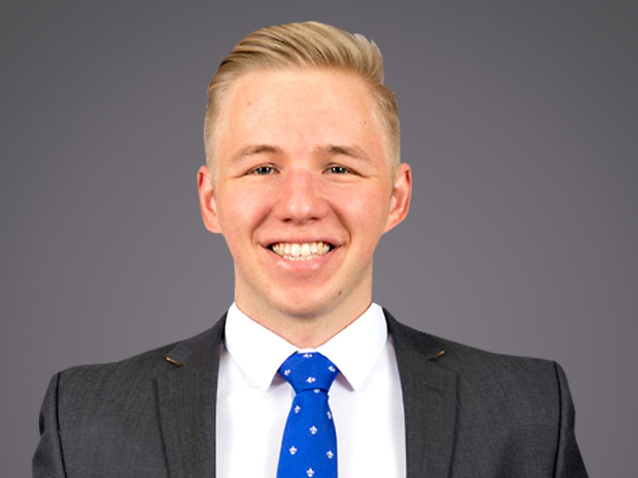 Portrait of a person with short blonde hair, wearing a dark grey suit, white shirt and blue patterned tie, smiling against a neutral grey studio background.