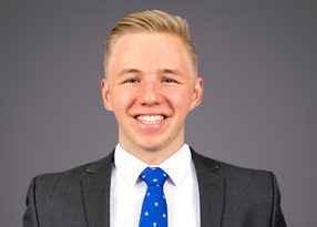 Portrait of a person with short blonde hair, wearing a dark grey suit, white shirt and blue patterned tie, smiling against a neutral grey studio background.