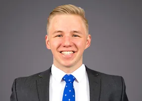Portrait of a person with short blonde hair, wearing a dark grey suit, white shirt and blue patterned tie, smiling against a neutral grey studio background.