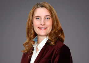 Portrait of a person with shoulder-length light brown hair, blue eyes, wearing a white shirt and burgundy blazer, smiling against a grey background.